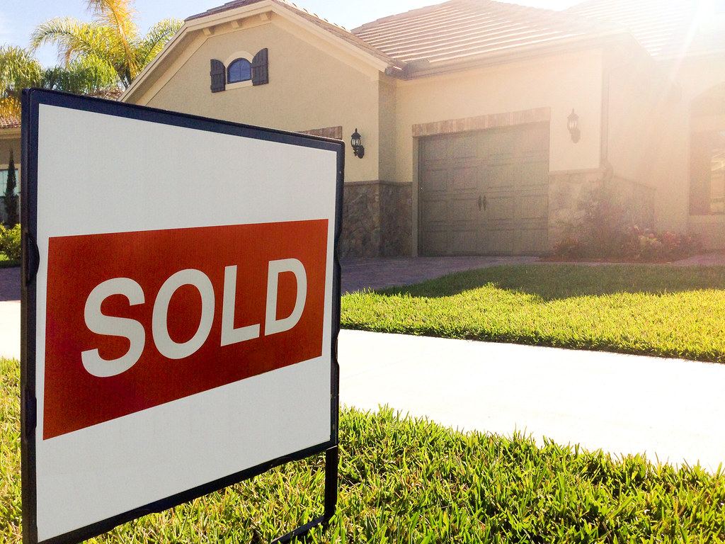 A red and white SOLD sign stands on a lawn in front of a stucco house with a garage and palm trees
