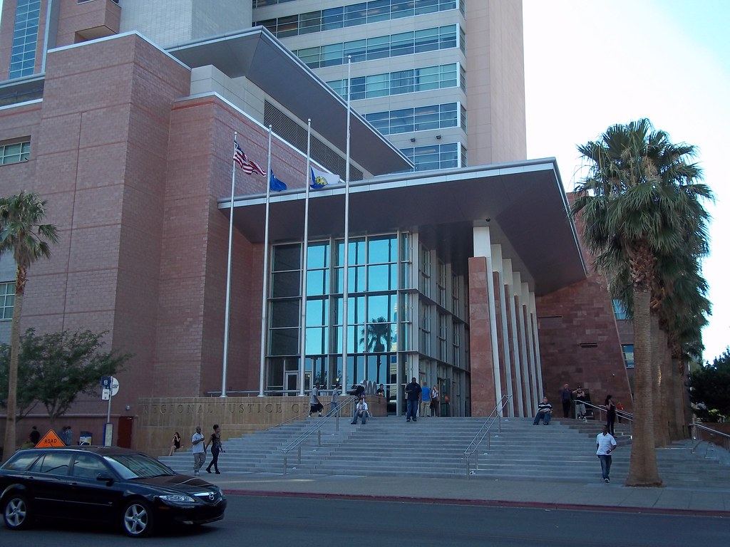 A large brick and glass courthouse with flags, wide front steps and palm trees, with a car passing on the street