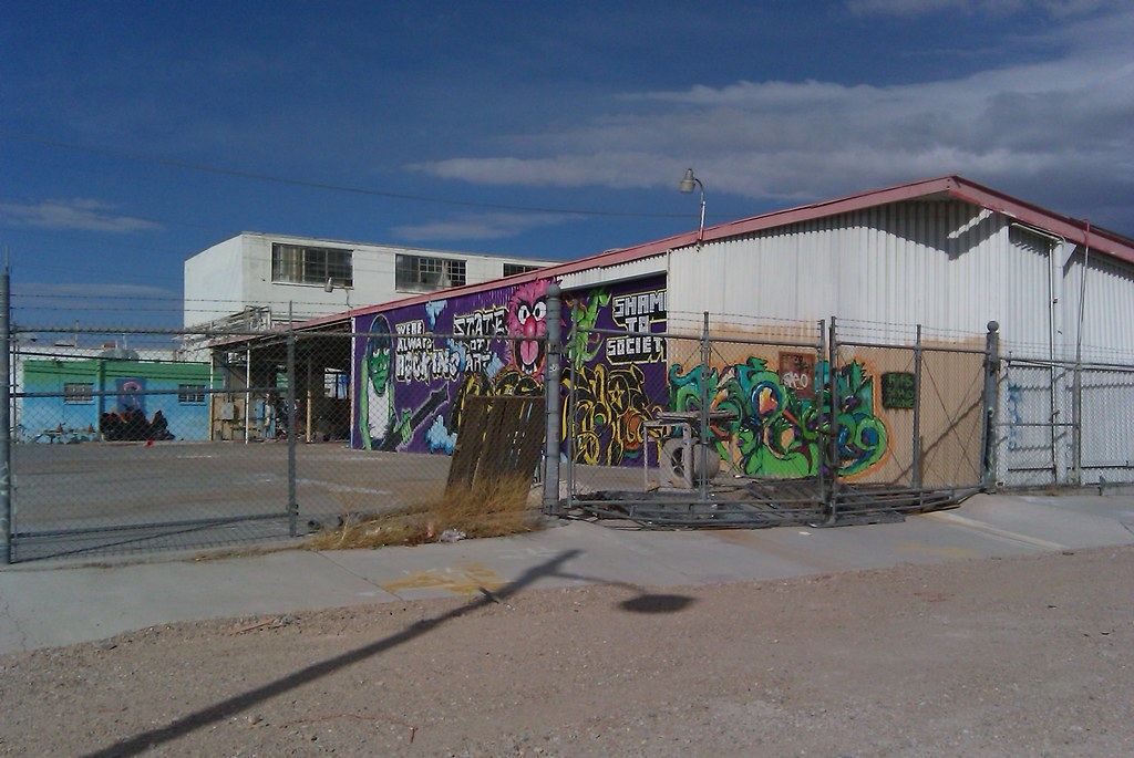A metal warehouse covered in colorful graffiti murals sits behind a chain-link fence beside an empty street