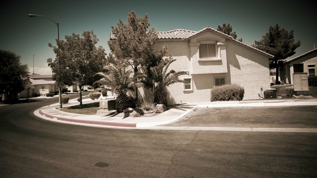 A two-story stucco house with a tile roof, palm trees and shrubs sits on a curved suburban street corner