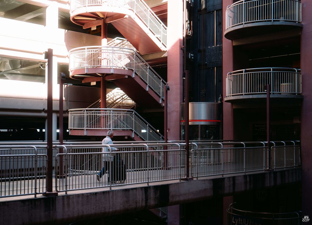A traveler pulls a suitcase across a walkway in a parking garage with red spiral staircases and railings