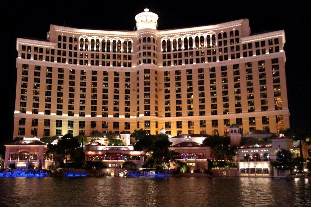 A large curved hotel tower lit up at night behind a lake, with lighted restaurants along the waterfront
