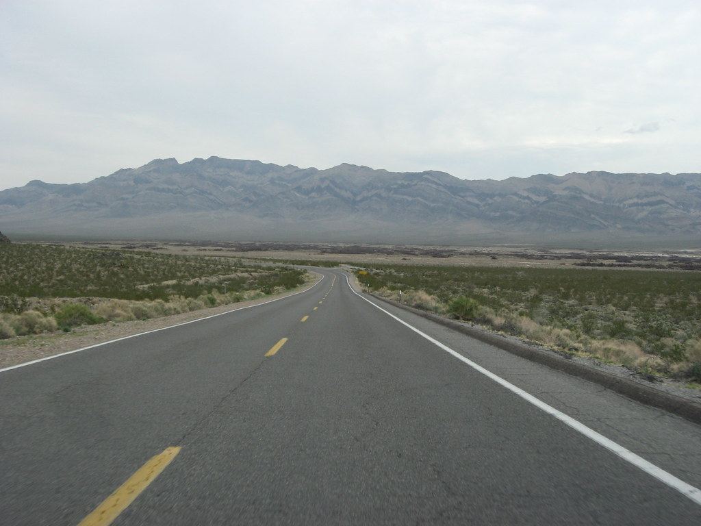 A two-lane desert highway curves through scrubland toward a rugged mountain range under an overcast sky