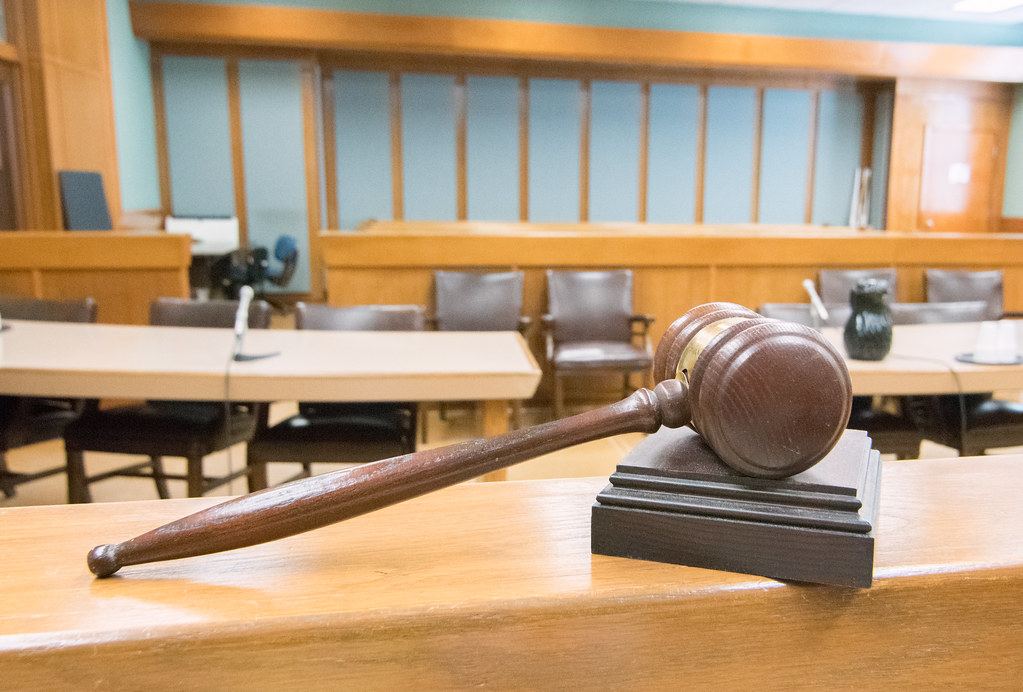 A wooden gavel rests on its block on a courtroom railing, with empty tables and chairs in the background