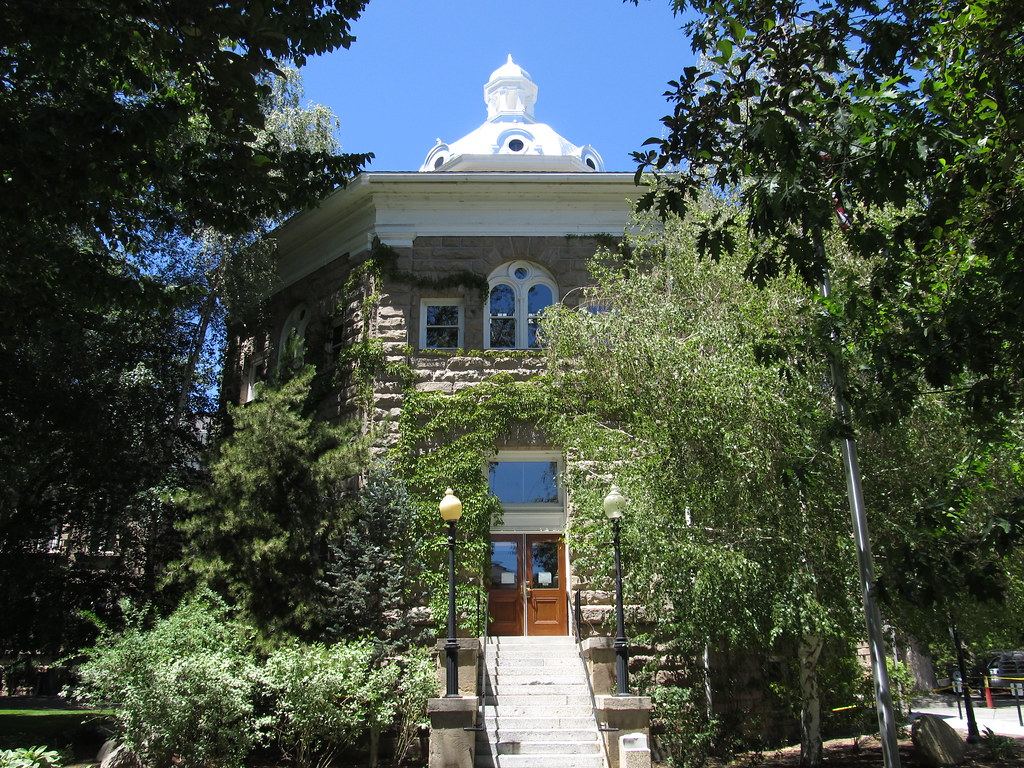 Ivy-covered sandstone building with a white cupola, stone steps and lampposts, framed by trees under a blue sky