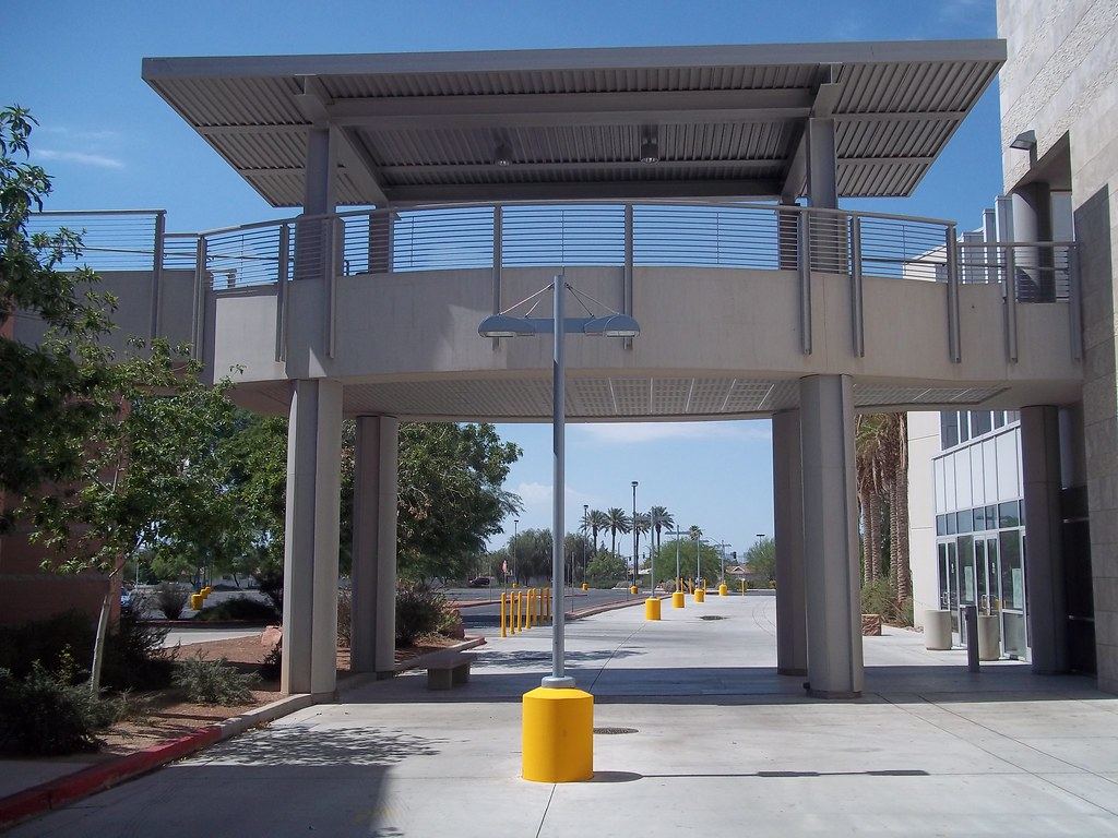 Covered concrete walkway and pedestrian bridge between campus buildings, with palm trees and yellow bollards