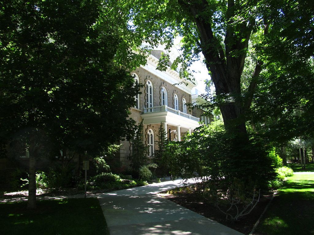 A sandstone building with arched windows and a white balcony, partly hidden by shade trees beside a walkway