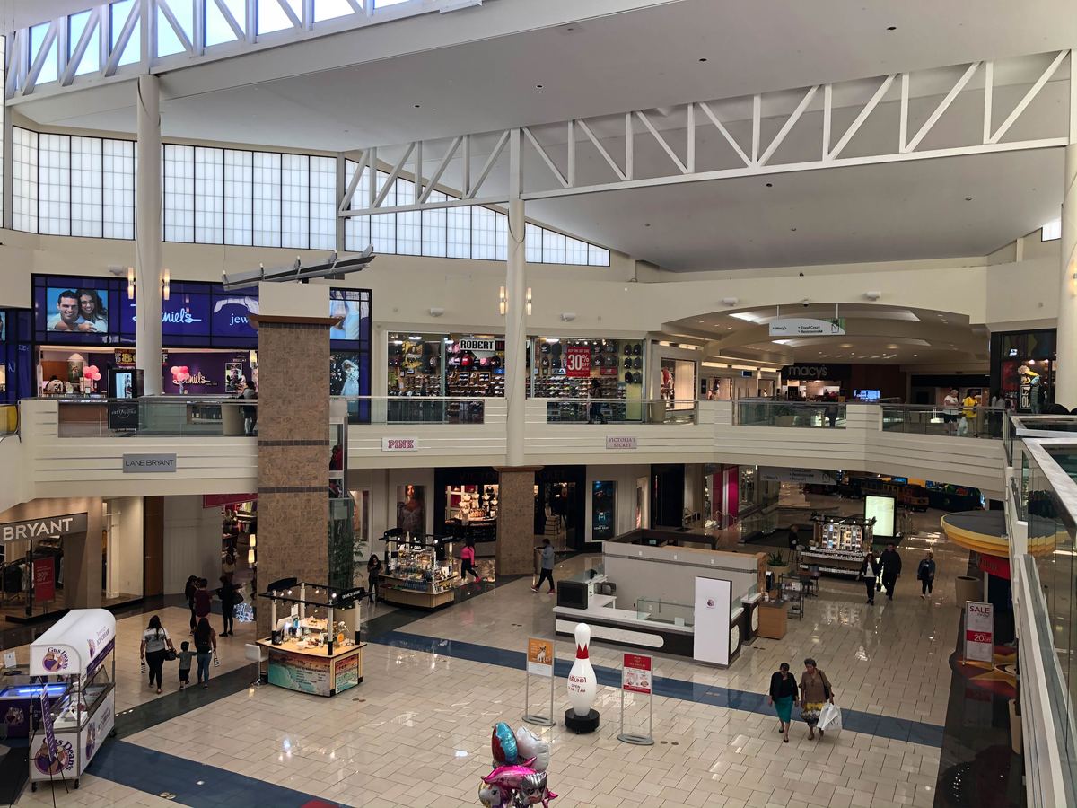 Two-level shopping mall interior with storefronts, kiosks, a skylit ceiling and a few shoppers walking