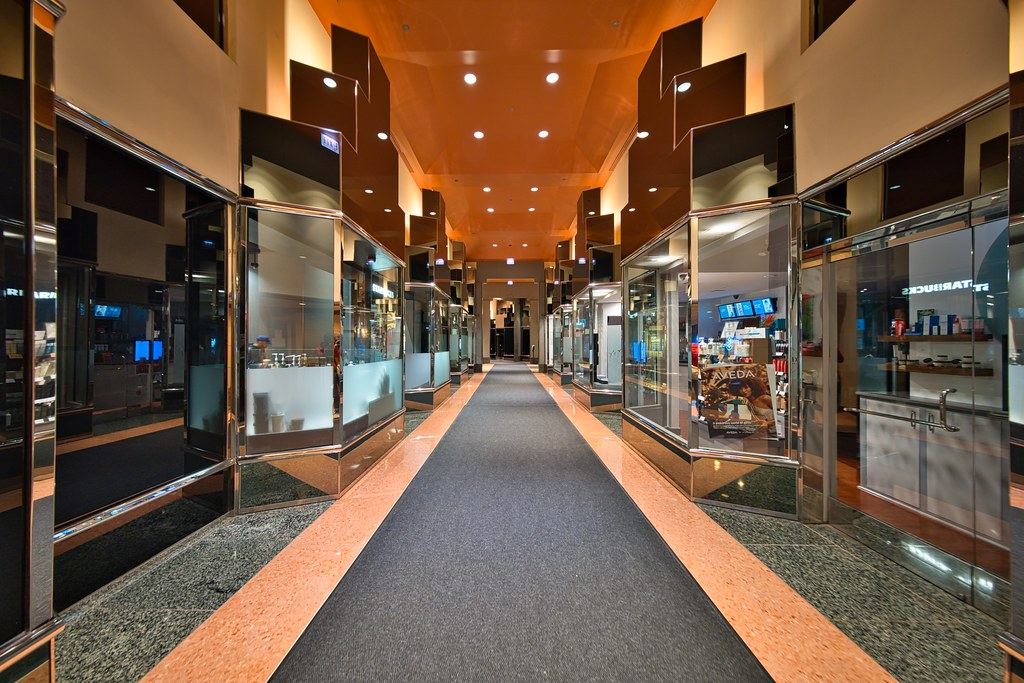 Empty indoor shopping mall corridor with a dark carpet runner, glass storefronts and orange-lit ceiling
