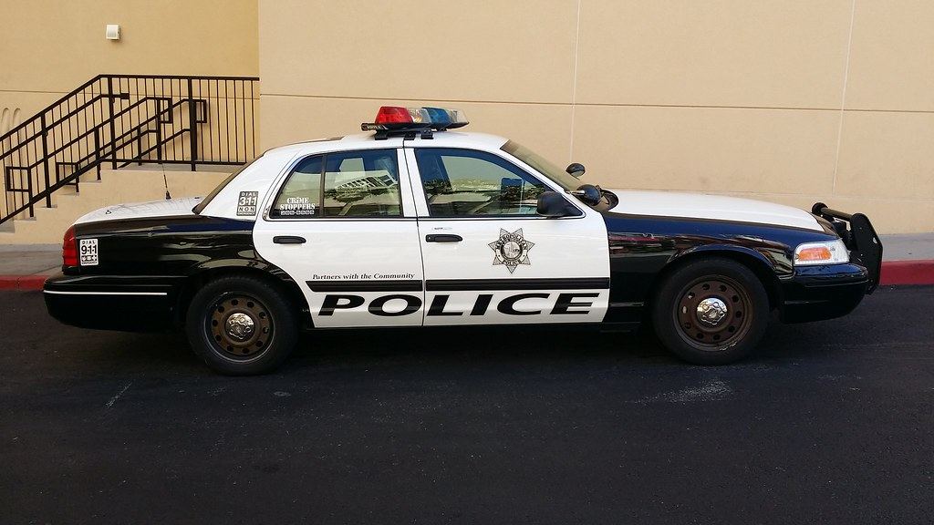 A black and white police sedan with a roof light bar parked beside a tan building and stairway