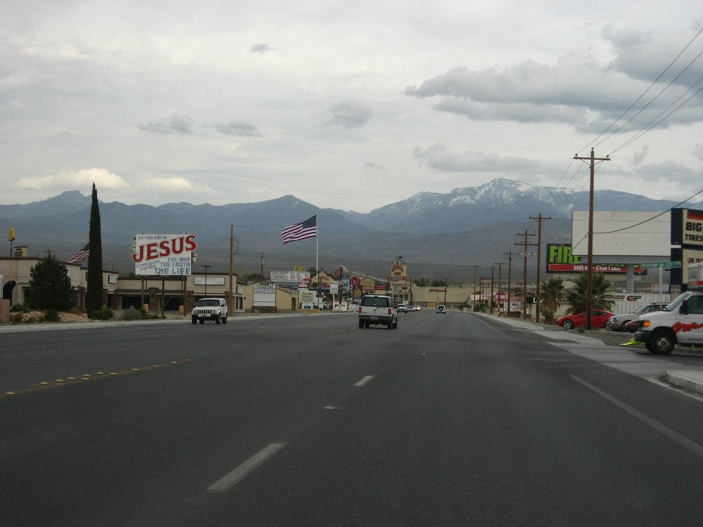 A wide desert town road lined with businesses, signs and an American flag, with mountains under a cloudy sky