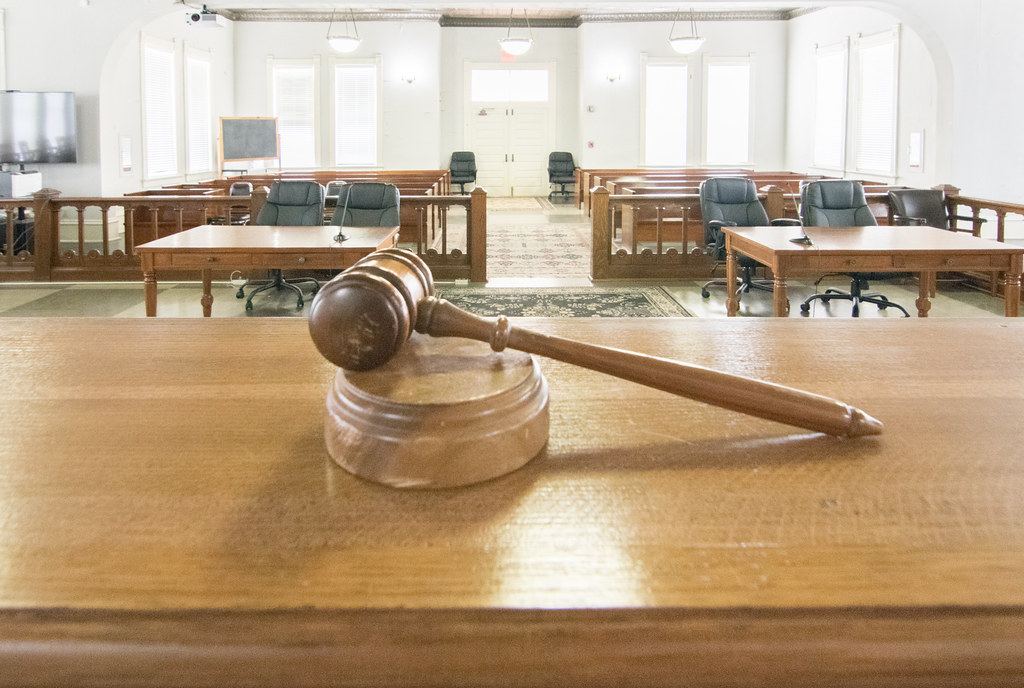 A wooden gavel rests on a judge's bench facing an empty courtroom with counsel tables and chairs