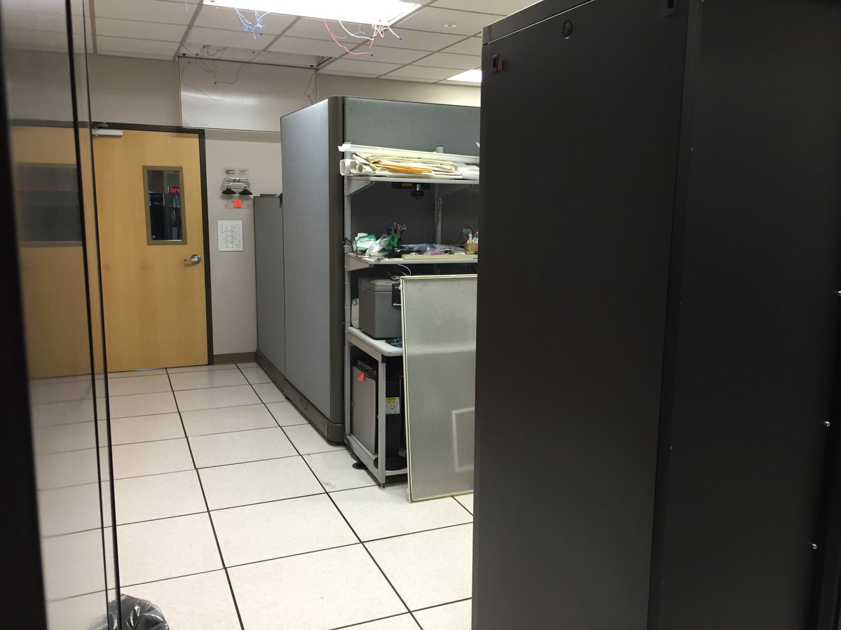 A server room with dark equipment racks, a raised white tile floor, a cluttered workstation and a wooden door