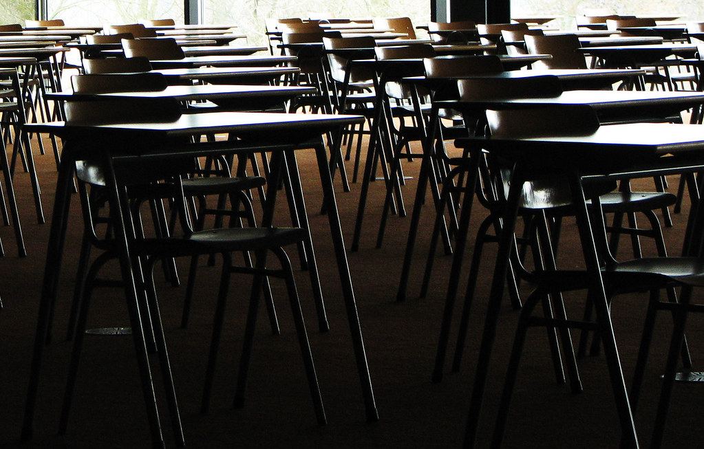 Rows of empty desks and chairs in a dim room lit by large windows