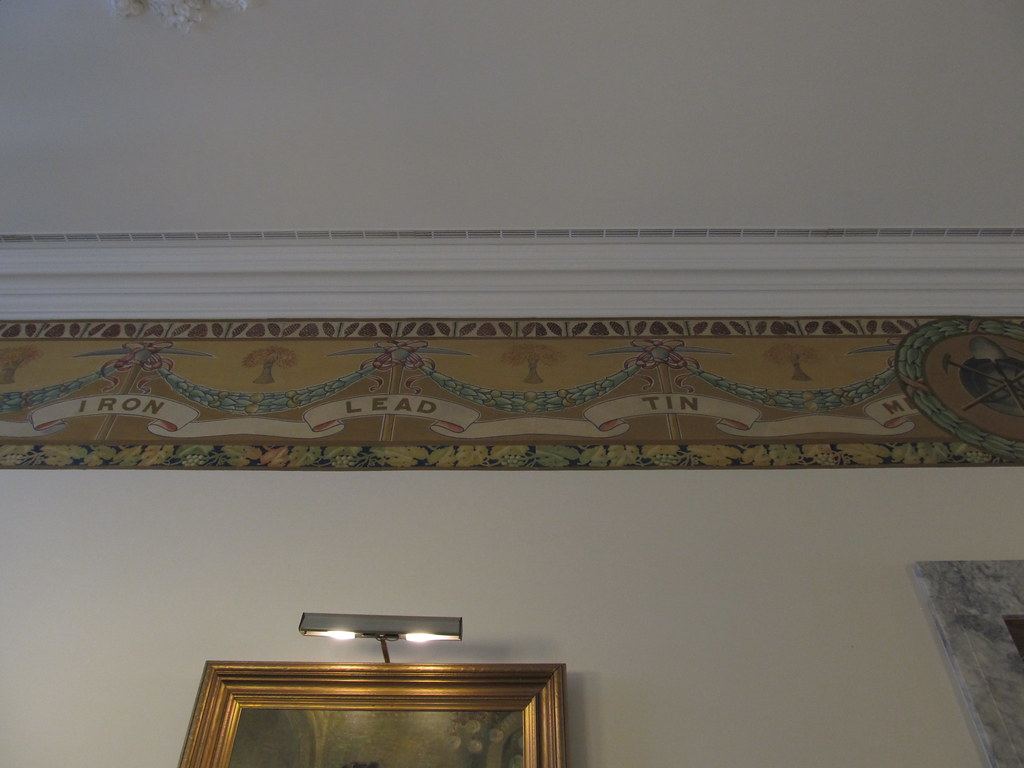 Decorative wall frieze with garlands and banners reading Iron, Lead and Tin above a framed painting lit by a lamp