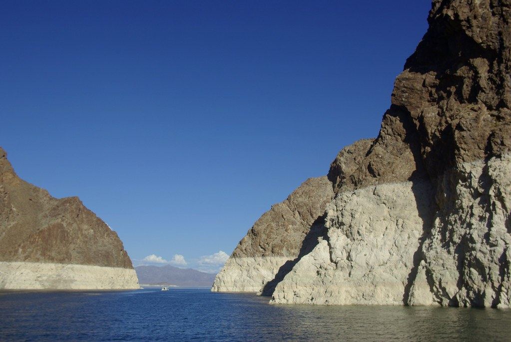 Rocky desert cliffs with a wide white mineral band above blue water, under a clear sky with a small boat in the distance