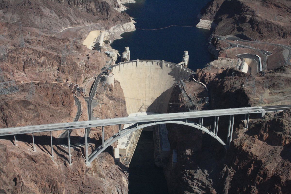 Aerial view of Hoover Dam holding back blue reservoir water, with an arched highway bridge spanning the rocky canyon