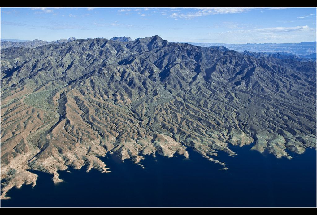Aerial view of rugged desert mountains sloping down to a deep blue lake with a pale band along the shoreline