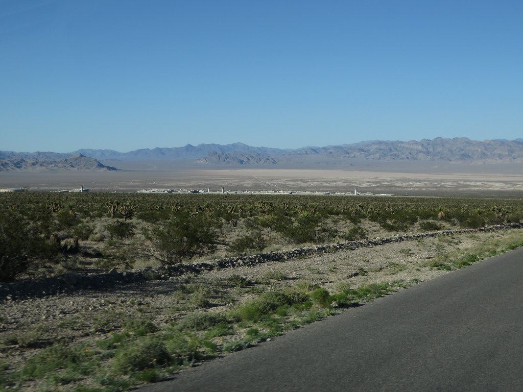 A distant prison complex with watchtowers sits in a desert valley below mountains, seen from a roadside