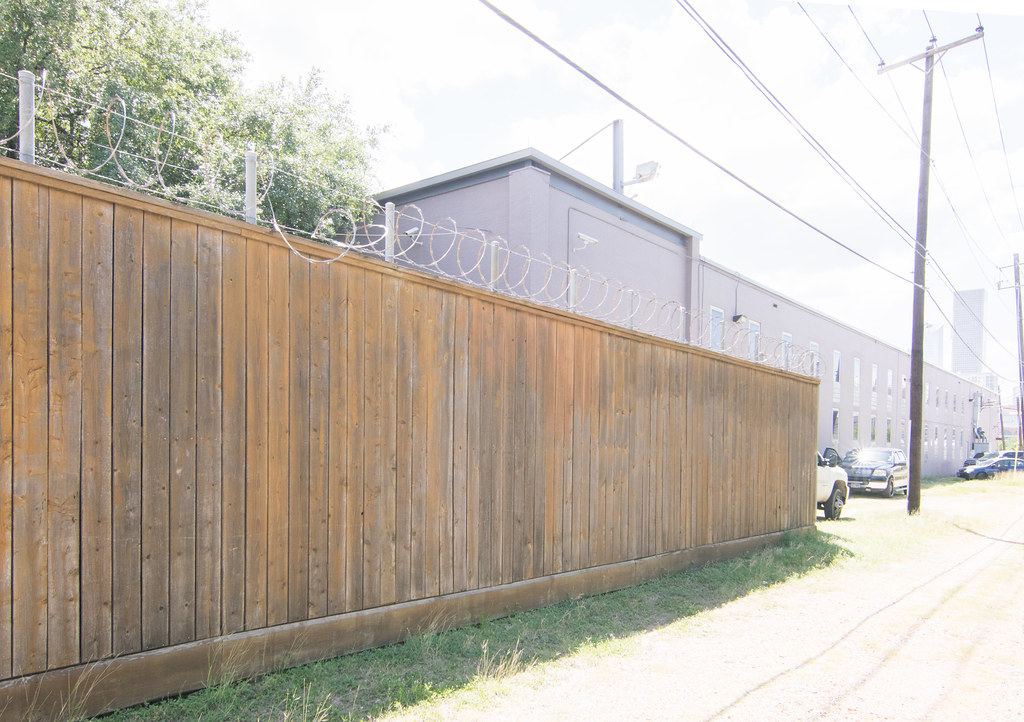 A tall wooden fence topped with coiled razor wire runs beside a gray building with security cameras