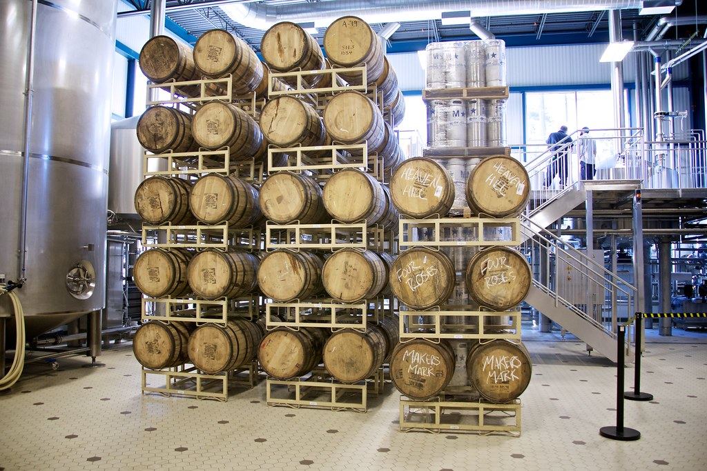 Wooden barrels stacked on metal racks beside steel tanks and a staircase inside a brightly lit production facility