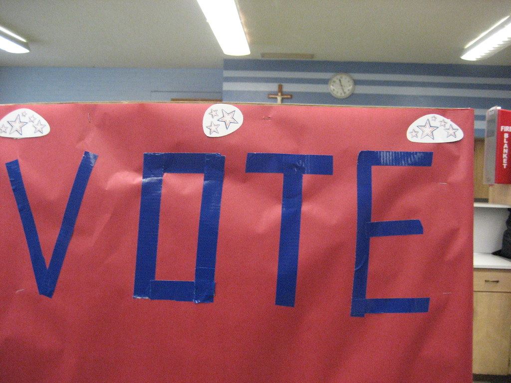 Red paper sign reading VOTE in blue tape with star decorations, set in a room with a clock and crucifix