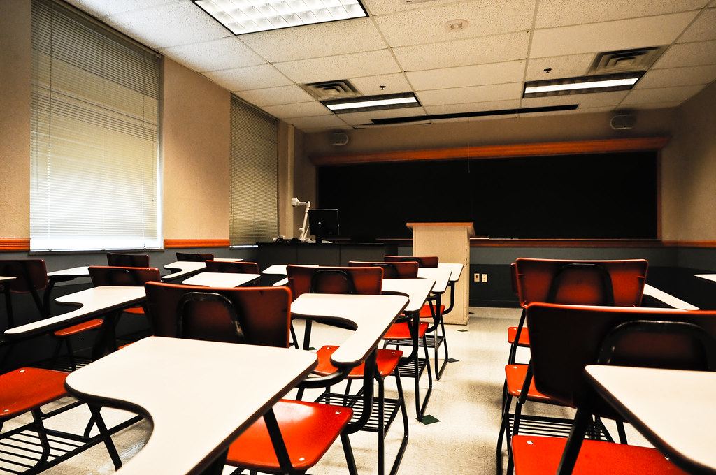 An empty classroom with rows of orange desk chairs facing a blackboard and a wooden lectern