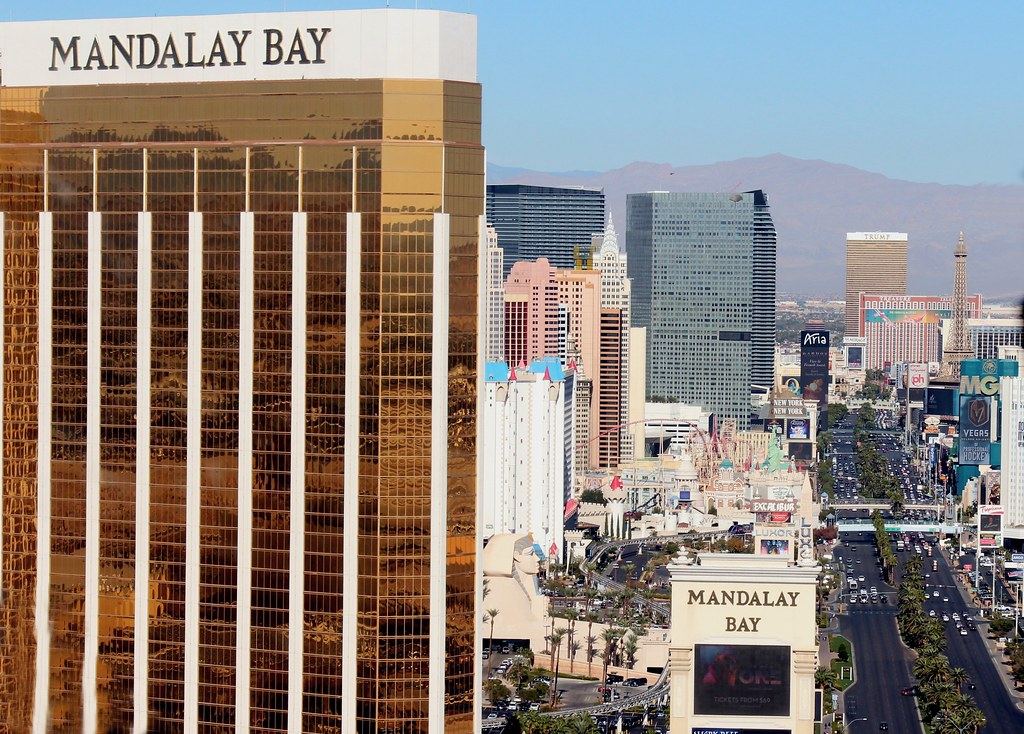 Gold Mandalay Bay tower beside Las Vegas Boulevard, lined with casino hotels and palm trees, mountains behind
