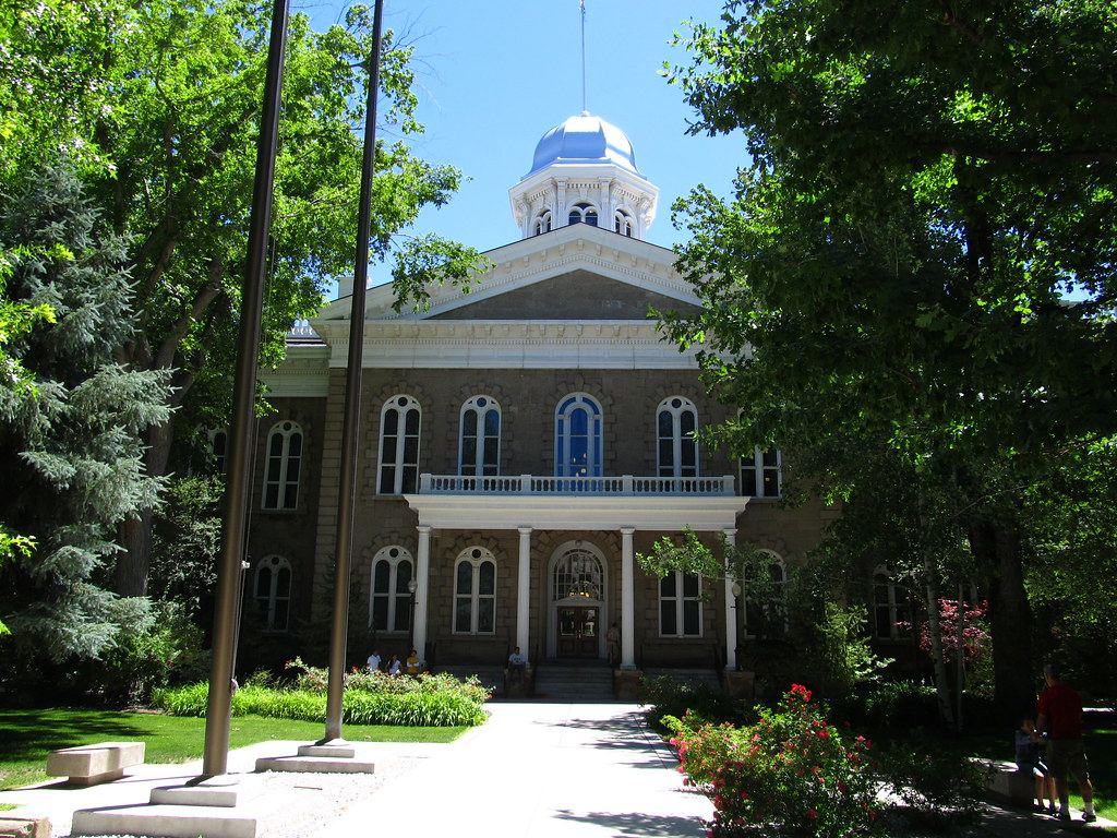 A sandstone capitol building with a silver dome and white columns, framed by green trees under a blue sky