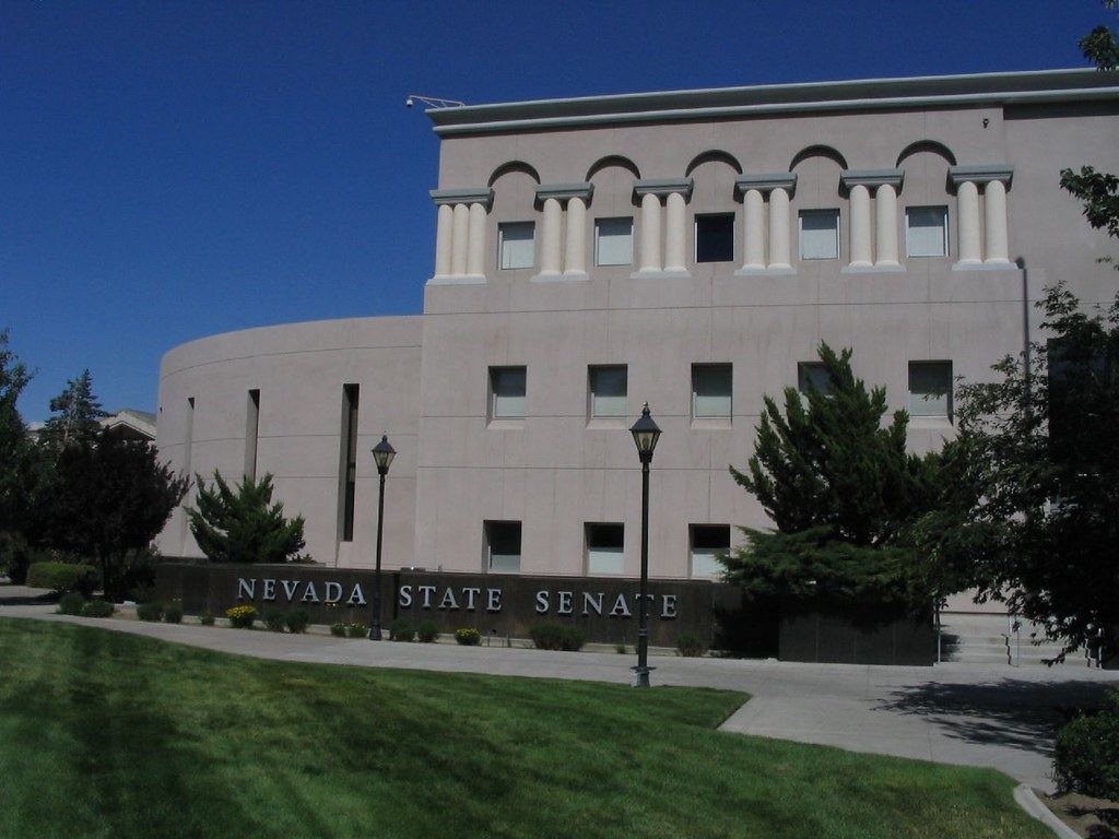 A pinkish-beige government building with columns and a sign reading Nevada State Senate, beside a green lawn
