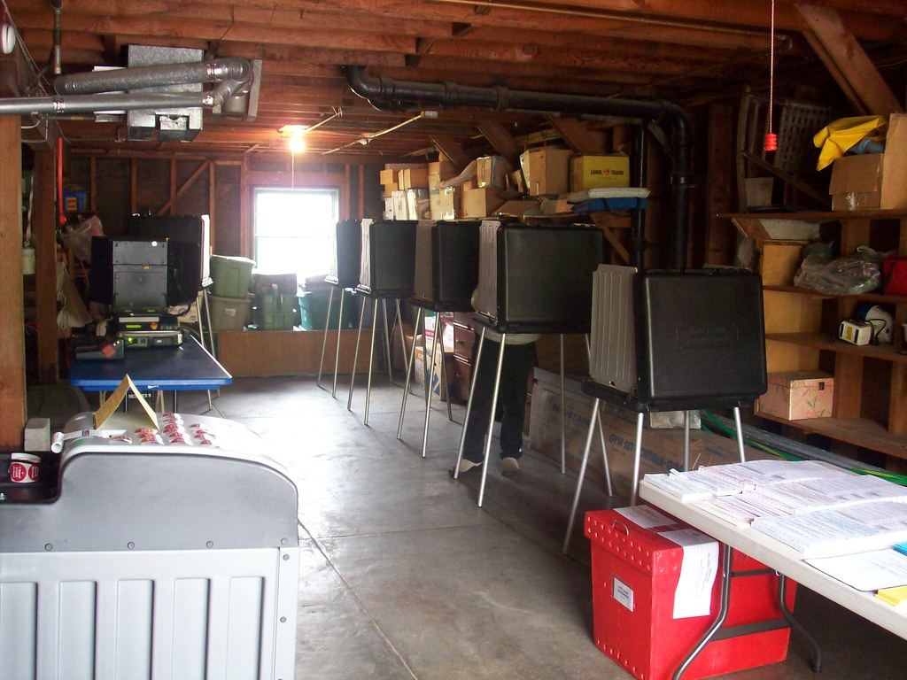 Row of portable voting booths in a garage, with a table of ballots and a red ballot box in the foreground