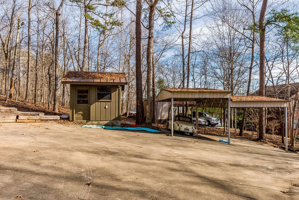 Metal carports sheltering a vintage car beside a small shed on a paved lot surrounded by bare trees