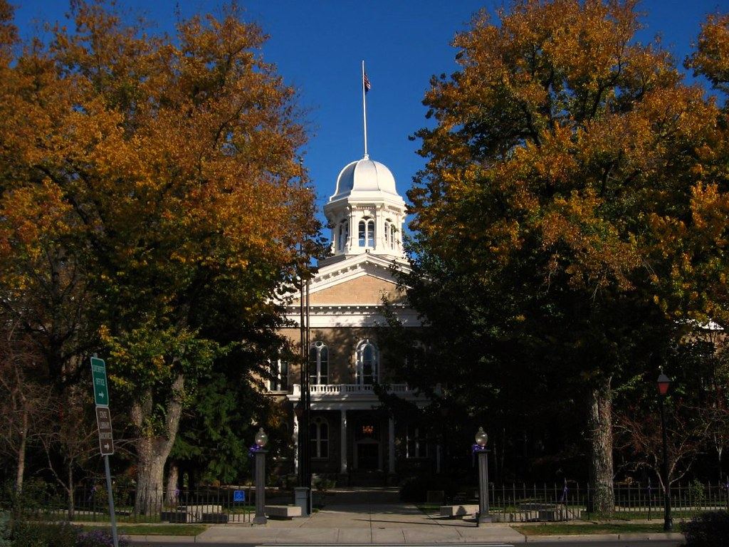 A domed sandstone capitol building with a flag on top, framed by autumn trees and an iron fence