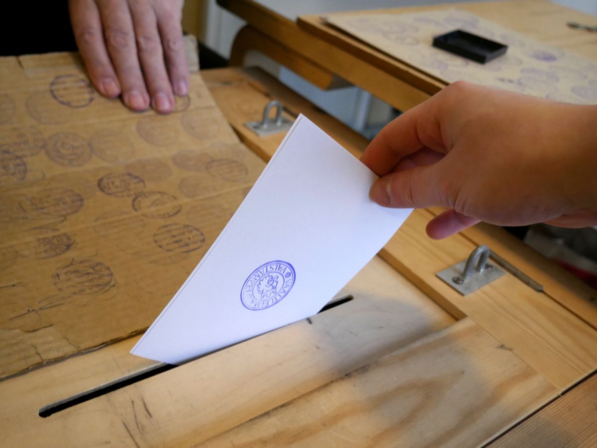 A hand drops a stamped white paper ballot into the slot of a wooden ballot box