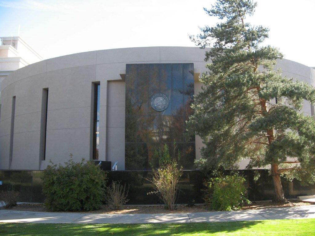 Curved beige government building with a dark glass panel bearing a state seal, beside a tall pine tree and shrubs