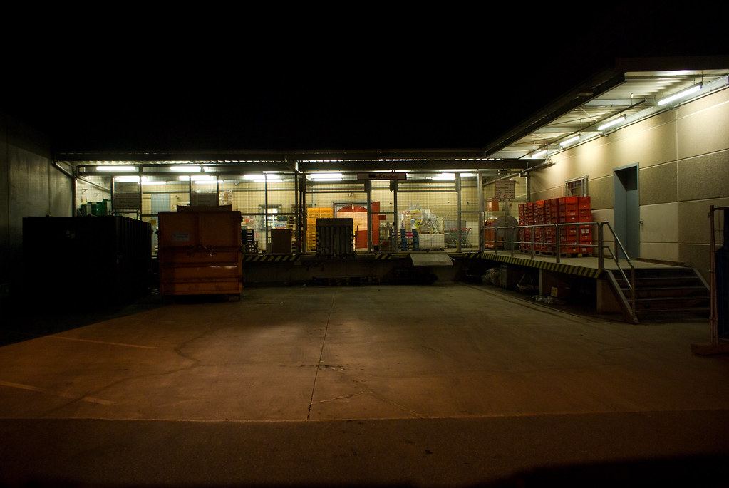 A lit warehouse loading dock at night with stacked red crates, dumpsters and an empty concrete yard