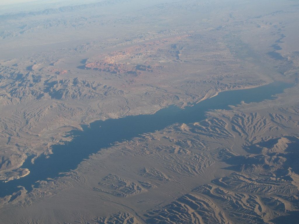 Aerial view of a long blue lake arm winding through dry, rugged desert mountains with red rock formations