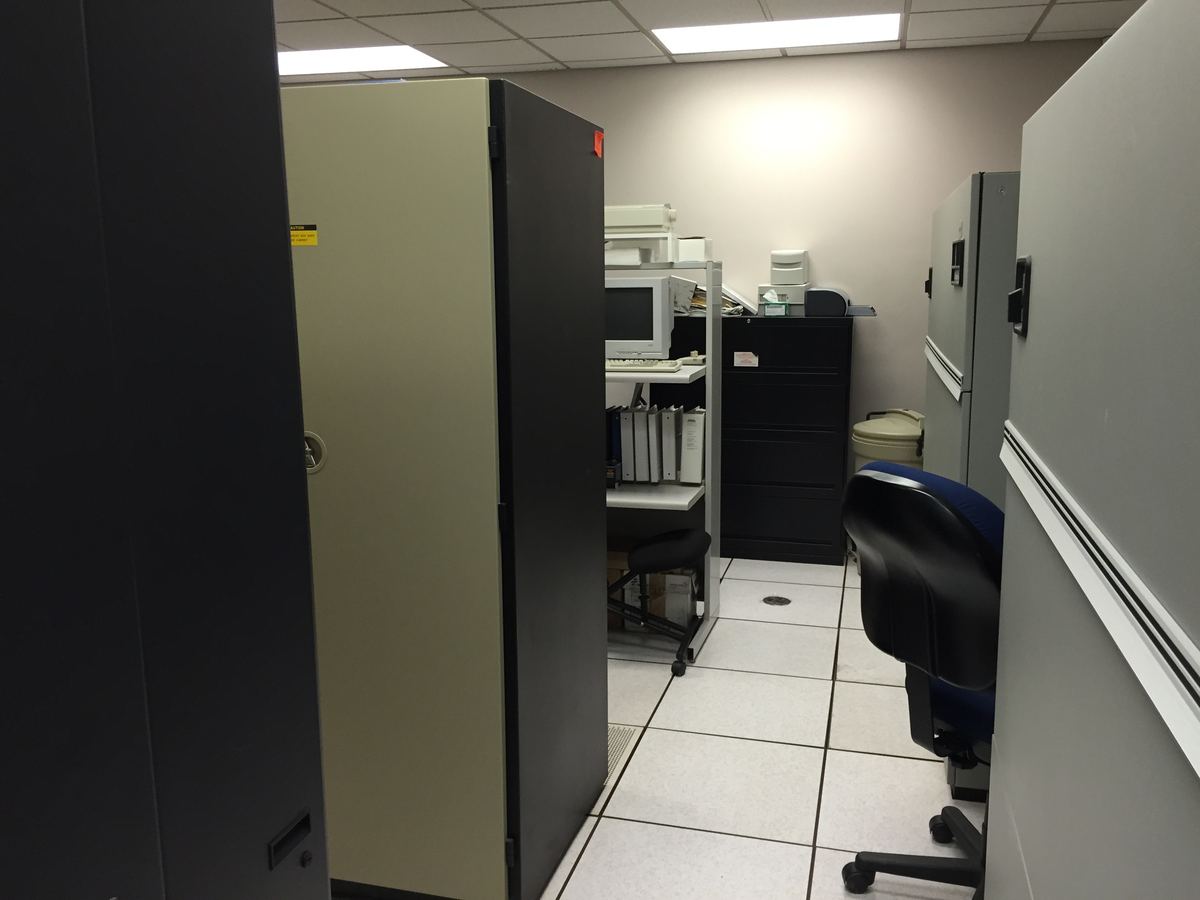 A small server room with tall equipment cabinets, a raised tile floor, an old monitor and an office chair