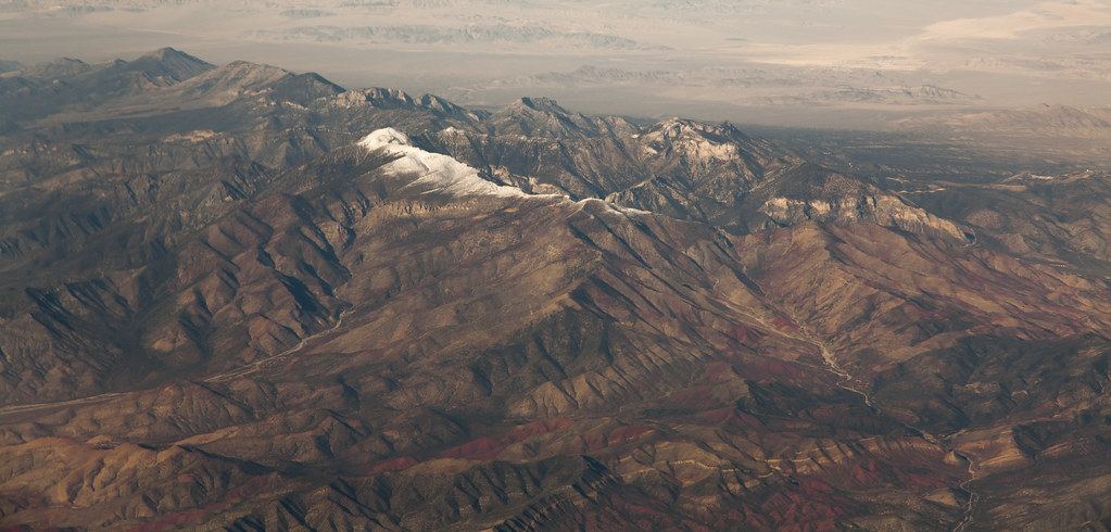 Aerial view of rugged brown desert mountains with a snow-dusted ridge and hazy basins beyond
