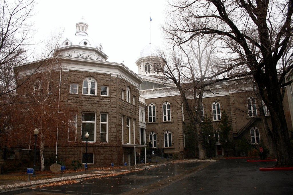 A sandstone capitol building with white domed cupolas behind bare trees on a gray, rainy day