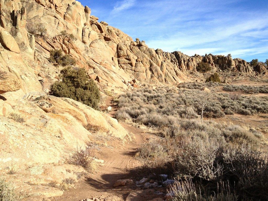 A narrow dirt trail winds past sagebrush and tan granite rock formations under a blue sky