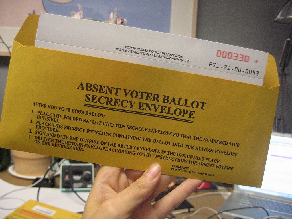 A hand holds a yellow absent voter ballot secrecy envelope with a numbered ballot stub inside