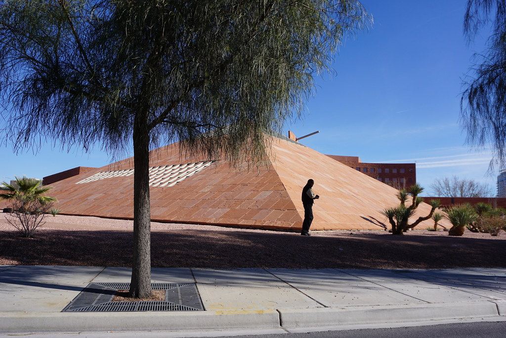 A red stone pyramid-shaped building behind a tree and desert plants under a clear blue sky