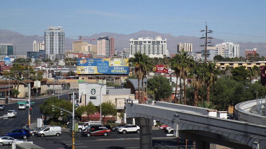 City skyline with high-rise towers and mountains behind, above a busy intersection, palm trees and an elevated track