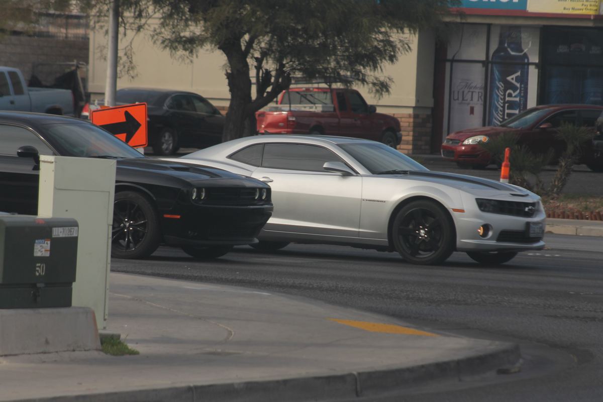 A silver Camaro and a black Challenger drive past a storefront and traffic cone on a city street