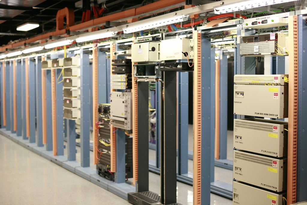 Rows of blue and orange equipment racks holding beige telecom and networking units in a brightly lit room