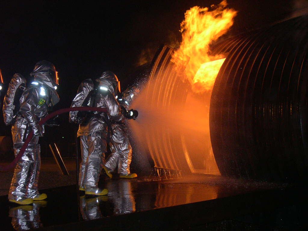 Firefighters in silver protective suits spray water at flames on a large metal training structure at night