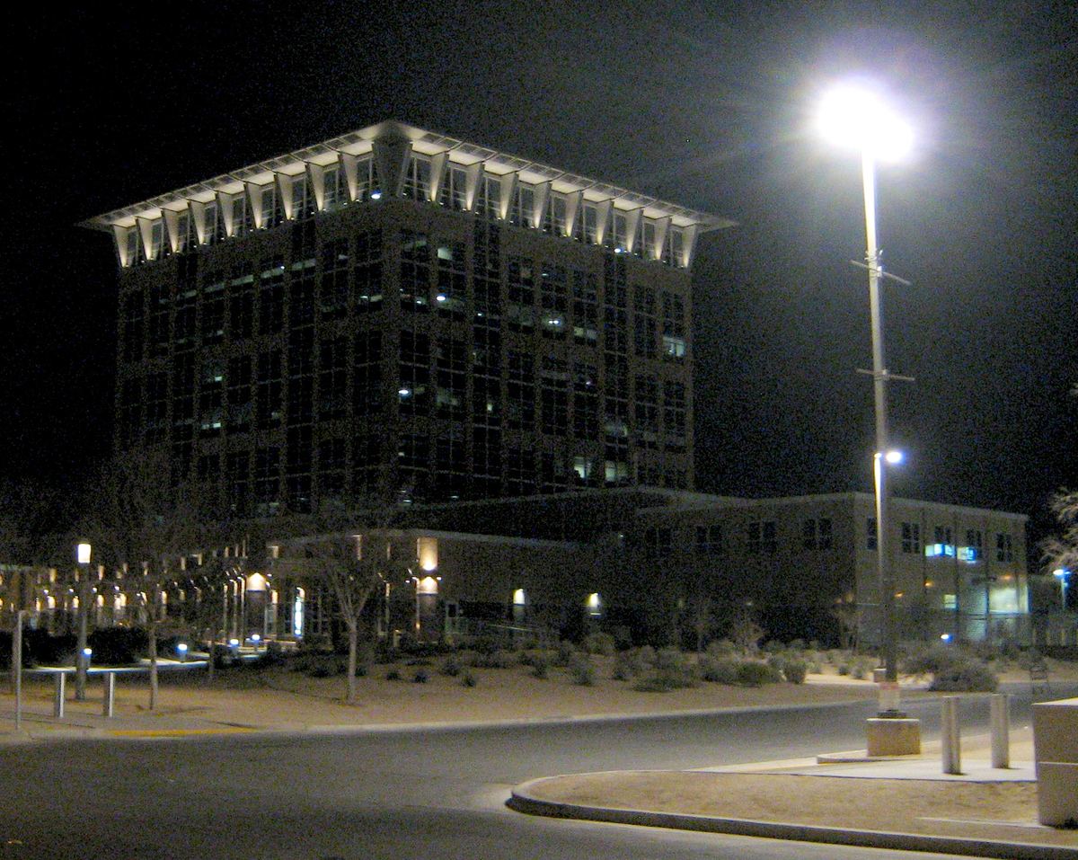 A multistory office building with an illuminated flared roofline at night, beside a lit street lamp and empty road