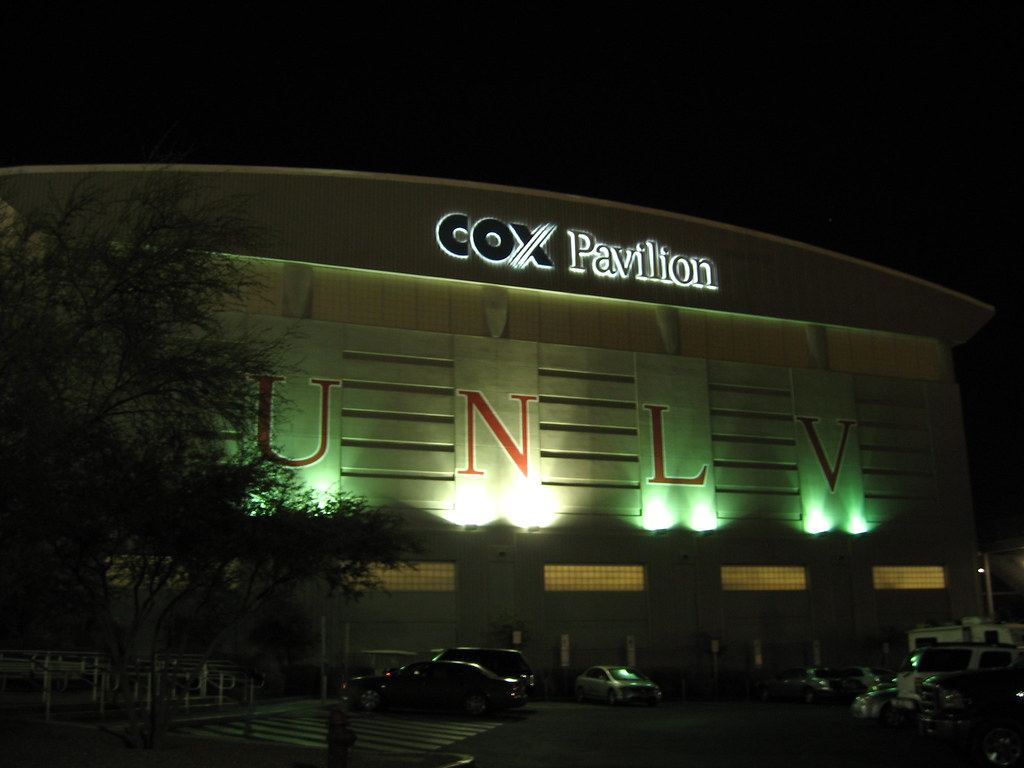 A large arena building lit up at night with UNLV letters and a Cox Pavilion sign above a parking lot