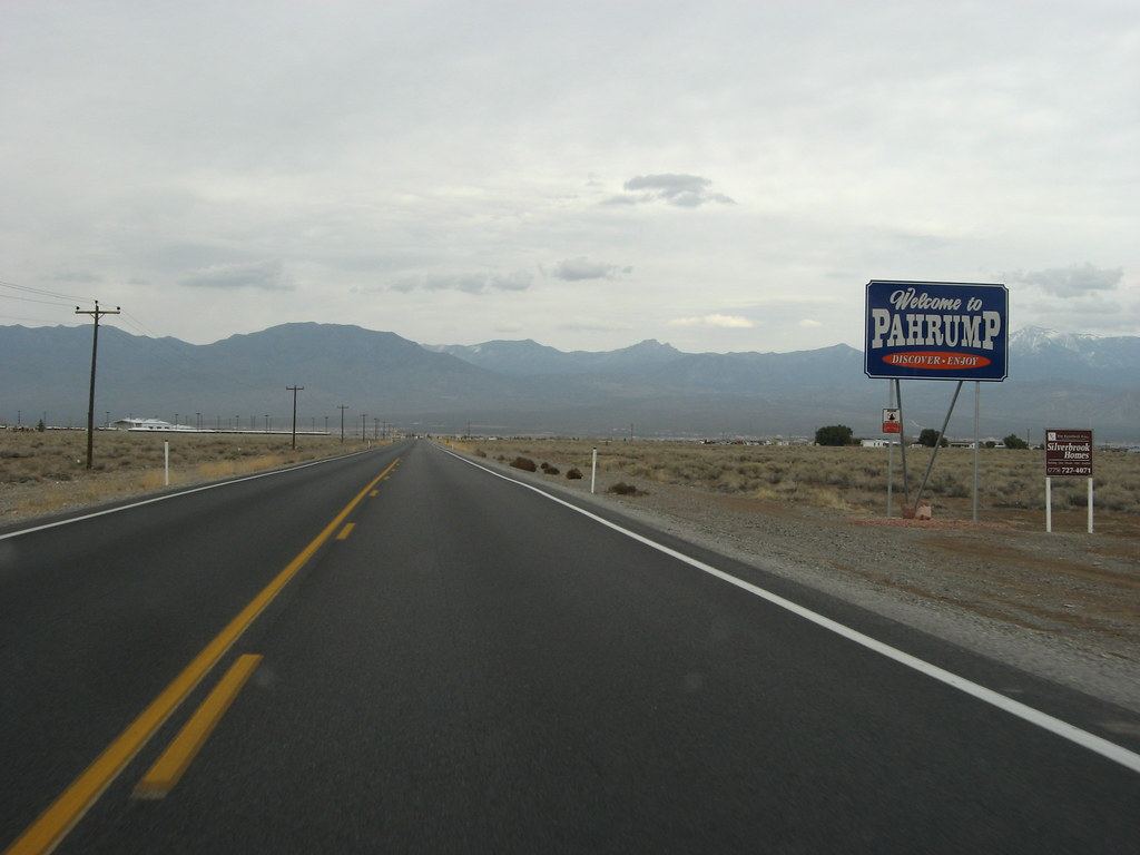 A desert highway passes a blue 'Welcome to Pahrump' sign with mountains under a cloudy sky